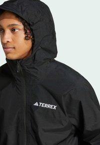 Sort vandtæt jakke med høj krave og justerbar hætte. Har en frontlynlås og et hvidt "TERREX"-logo på brystet.