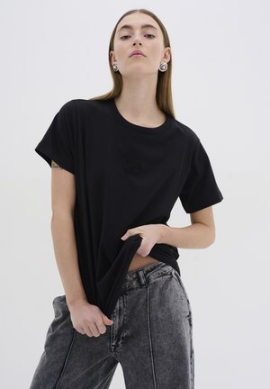 T-shirt in cotone nero con scollatura rotonda e maniche corte, con un logo goffrato discreto sul fronte. Indossato con jeans grigi strappati.