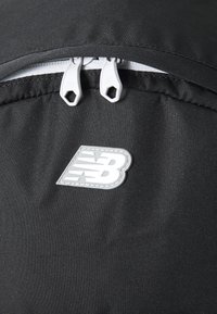 Saco de tecido preto com puxadores duplos de zíper cinza e um logótipo da New Balance em prata. O material tem uma textura suave com um brilho subtil.