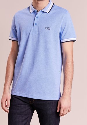 Muž oblečený v světle modré polokošili Hugo Boss s černým a bílým lemováním na límci a rukávech, v kombinaci s tmavými džínami.