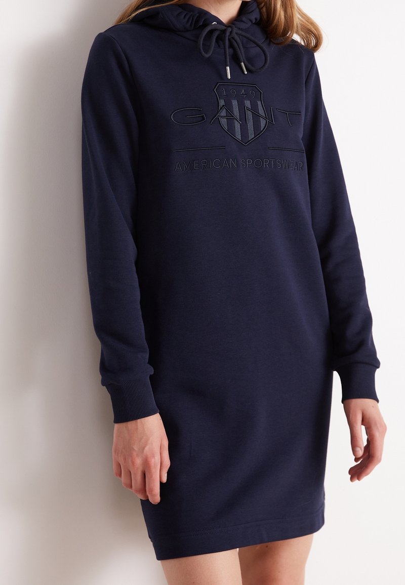 Femme portant une robe à capuche bleu marine à manches longues avec le logo GANT et le texte "American Sportswear" brodé sur le devant.