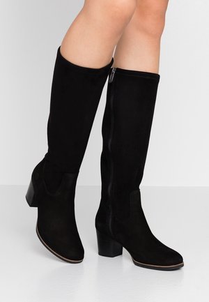 Czarne botki za kostkę na słupku z bocznymi zamkami, noszone na gołych nogach na jednolitym jasnym tle.