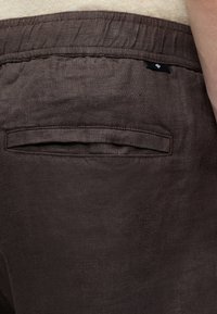 Gros plan d’un pantalon en lin marron avec une taille élastique et une poche passepoilée arrière porté par une personne sur un fond clair.