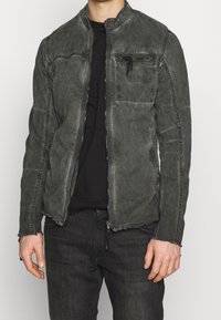 Man som bär en urtvättad svart zip-up jacka med synlig sömnad och en zip-fick på bröstet, tillsammans med svarta jeans och en svart skjorta.