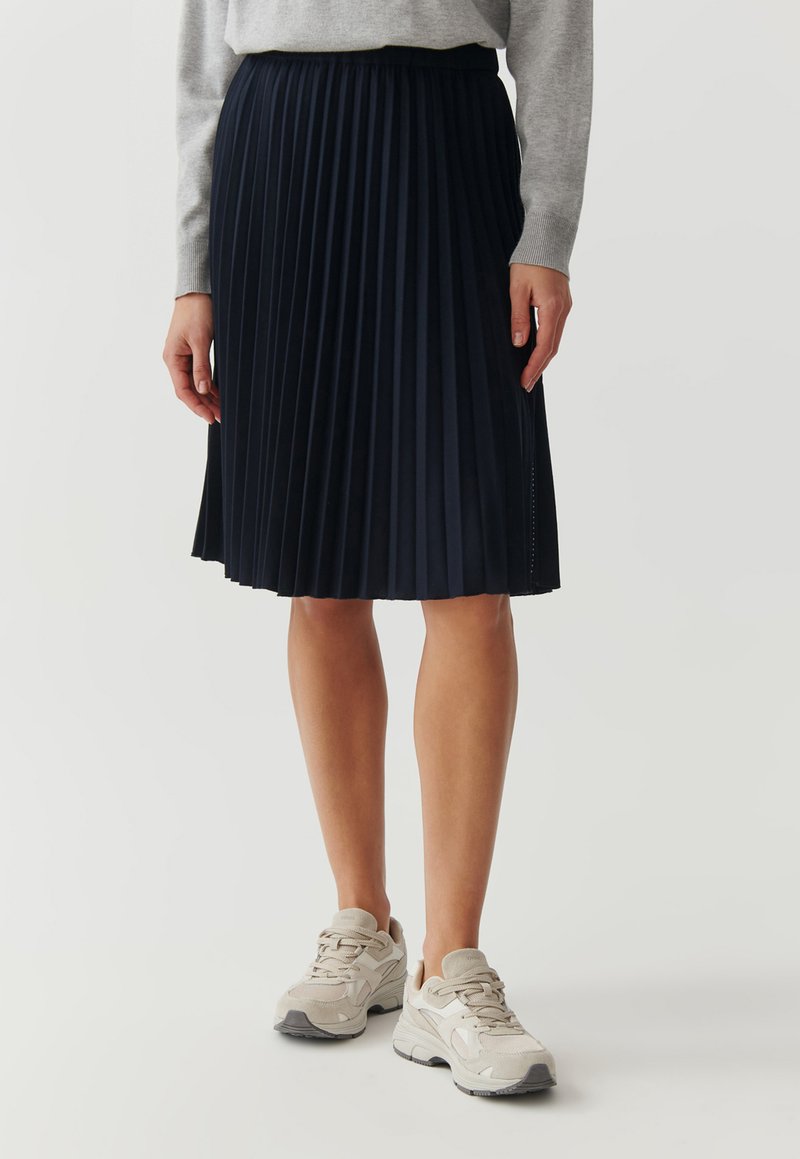 Femme portant une jupe plissée bleu marine jusqu'aux genoux, un haut à manches longues gris et des baskets chunky beige, se tenant sur un fond blanc uni.
