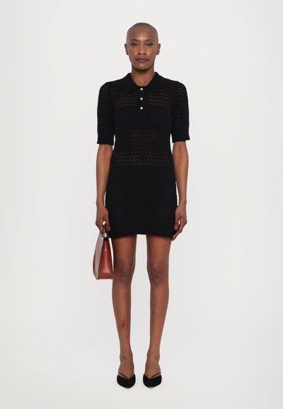 Schwarzes gestricktes Polo-Kleid mit kurzen Ärmeln, mit einem geknöpften Kragen, strukturiertem Muster und einer figurbetonten Silhouette. Das Model hält eine braune Handtasche.