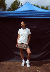 Biela textúrovaná košeľa Nike s modrým golierom a červeným logom, v kombinácii s béžovou sukňou. Model má na sebe šedé tenisky a biele ponožky, stojí vonku.