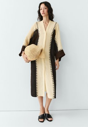 Donna che indossa un lungo vestito beige e marrone scuro, che tiene un cappello di paglia intrecciato, in piedi davanti a uno sfondo bianco semplice.