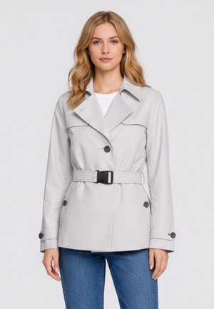 Giovane donna con capelli biondi che indossa una giacca grigio chiaro con cintura e jeans blu, in piedi davanti a uno sfondo bianco uniforme.