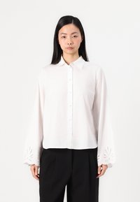 Hvit skjorte med krage, lange ermer som har broderte utskjæringer ved mansjettene, laget av lett stoff.