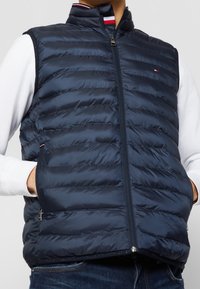 Granatowa pikowana kamizelka z wysokim kołnierzem, zamkiem błyskawicznym, pikowaną teksturą oraz bocznymi kieszeniami. Kołnierz ozdobiony czerwono-białymi paskami.