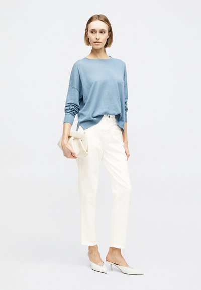 Femme portant un pull à manches longues bleu clair, un pantalon blanc taille haute, des mules blanches à talons pointus, tenant une pochette beige.