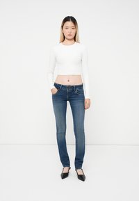 Una mujer joven está de pie frente a la cámara, vistiendo una camiseta blanca de manga larga, jeans azules y zapatos de punta negros, con un fondo blanco liso.
