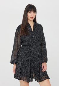 Abito nero con pois bianchi, bottoni frontali, colletto, maniche lunghe trasparenti, cintura in vita e gonna svasata con una leggera apertura nella parte inferiore.