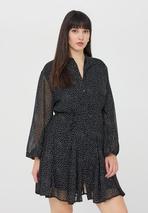 Abito nero con pois bianchi, bottoni frontali, colletto, maniche lunghe trasparenti, cintura in vita e gonna svasata con una leggera apertura nella parte inferiore.