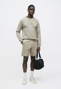 Fiatal férfi bézs EA7 pulóverben és hozzáillő rövidnadrágban, fehér zokniban és sportcipőben, fekete válltáskát tartva egyszerű háttér előtt.