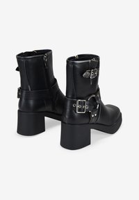 Botas de cuero negro hasta el tobillo con tacones gruesos, hebillas plateadas y cremalleras; textura suave; correas aseguradas alrededor del tobillo.