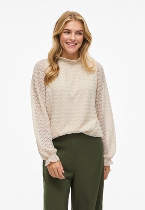 Beige blouse with a textured, zigzag pattern, long sheer sleeves, and a ruffled neckline. Paired with dark green trousers.