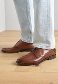 Braune Lederanzugschuhe mit glatter Oberfläche, spitzem Zehenbereich und dezentem Schnürsenkel, kombiniert mit hellblauen Denim-Hosen auf einem Holzboden.