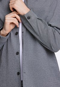 Graues Button-Up-Hemd aus strukturiertem Stoff, ausgestattet mit schwarzen Knöpfen und einem lässigen Schnitt, unter dem ein sichtbares weißes Unterhemd hervorblitzt.