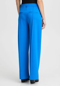 Spilgtas zilas platajās bikses ar gludu tekstūru, kurām ir divi aizmugurējie kabatas un pieguloša josta. Valkātas ar melnām kurpēm.