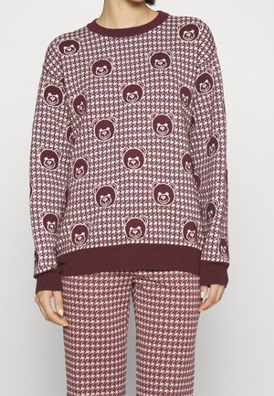 Personne portant un pull et un pantalon assortis à motif pied-de-poule bordeaux et rose avec des motifs répétés de visage d’ours sur le pull.