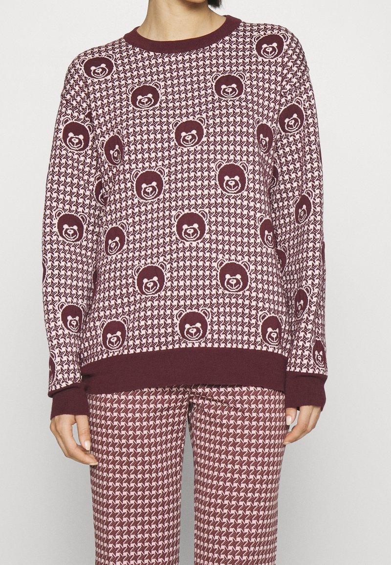 Personne portant un pull et un pantalon assortis à motif pied-de-poule bordeaux et rose avec des motifs répétés de visage d’ours sur le pull.