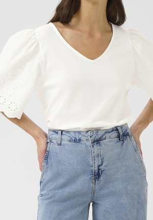 Kvinde iført hvid V-hals bluse med pufærmer og hulmønsterdetaljer samt blå højtaljede denimjeans med hænderne hvilende på hofterne.