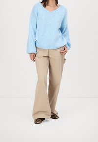 Femme portant un pull ample bleu clair, un pantalon large beige et des chaussures à bout pointu imprimé léopard, debout sur fond blanc.