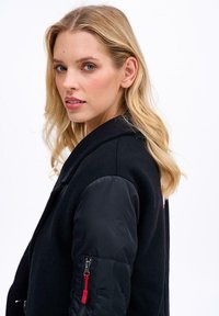 Blondýna s modrýma očima, oblečená v černém kabátu s prošívanými rukávy a kapsou na zip s červeným jezdcem, hledící přes rameno.
