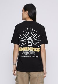 Isik, kellel on pikad mustad juuksed, kannab musta T-särki, mille seljal on valge kohvijänku graafika ja tekst "Laisk Pühapäeva Kohvi Klubi".