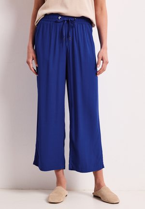 Kvinde iført løse, blå bukser med vide ben og snøre i taljen samt beige slip-on sko, stående op ad en ensfarvet lys væg.