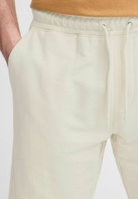 Un pantalon de survêtement beige clair avec une taille élastique et un cordon de serrage, doté de poches latérales et d'une texture de tissu douce et lisse.