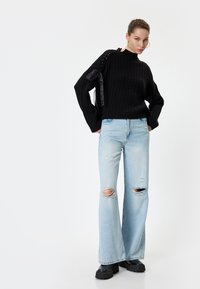 Zwarte geribbelde trui met een hoge hals en zijstrikdetails, gecombineerd met lichtblauwe, gescheurde, wijde jeans en flinke zwarte laarzen.