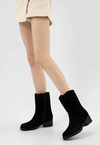 Gambe che indossano pantaloni corti beige e stivali neri in suede fino al polpaccio con tacco basso su uno sfondo bianco uniforme.