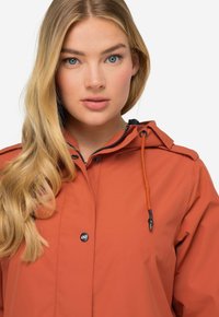 Oranssi vesitiivis takki hupulla, nepparikiinnitys, mustat vetoketjuyksityiskohdat ja säädettävä kiristysnauha kaulassa. Sileä kankaan pinta.