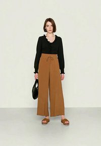 Blusa preta de manga longa com decote franzido, combinada com calças largas castanhas e sandálias de plataforma castanhas. Mochila preta segura na mão esquerda.