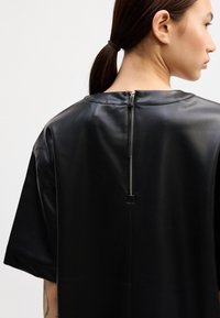 Vestido de couro preto com um corte descontraído. Apresenta um decote redondo e um detalhe de fecho vertical nas costas. Textura suave, design minimalista.