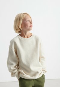 Krämsfärgad fleece-tröja med rund halsringning och sänkta axelsömmar, kombinerad med gröna byxor. Slät textur, avslappnad passform.