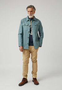 Chaqueta azul claro con dos bolsillos en el pecho y botones, llevada sobre una camiseta gráfica azul marino. Pantalones beige y botas marrones con cordones completan el conjunto.