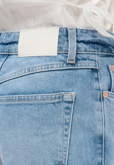 Jean en denim bleu clair avec une poche arrière, des rivets en cuivre et une étiquette de marque en cuir blanc sur la ceinture, porté avec un haut blanc texturé.