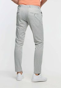 En man som bär ljusgrå slim-fit byxor och vita sneakers, står mot en vanlig vit bakgrund.