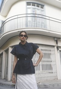 Crna košulja kratkih rukava s remenom u struku, srebrnim detaljima u obliku okova, u kombinaciji s teksturiranom bijelom suknjom. Model nosi crne sunčane naočale.