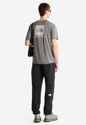 Mann mit Sonnenbrille, grauem The North Face T-Shirt, schwarzen Hosen mit Logo, trägt schwarze Umhängetasche, steht mit dem Rücken zur Kamera.