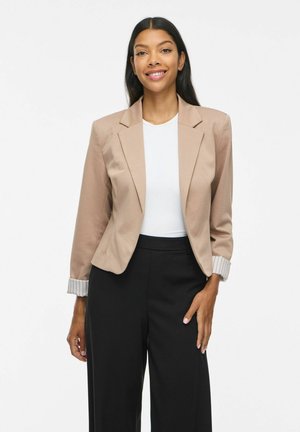 Donna sorridente con lunghi capelli neri indossa un blazer beige, una maglietta bianca e pantaloni neri, in piedi davanti a uno sfondo bianco semplice.