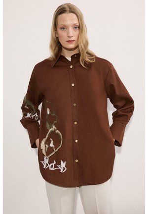 Kobieta o długich blond włosach, ubrana w brązową koszulę zapinaną na guziki z kwiatowym haftem na rękawach i przodzie, w połączeniu z białymi spodniami.