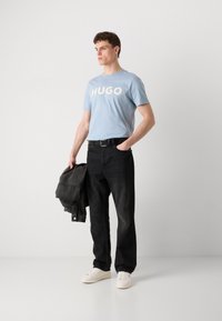 Világoskék pamut póló "HUGO" felirattal, fekete farmernadrág, fehér sportcipő, sötét szürke denim dzsekit tartva, egy egyszínű háttér előtt áll.