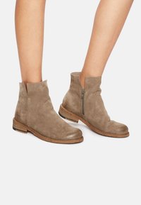 Stivali ankle in suede taupe con punta rotonda, chiusura con zip laterale e tacco basso a blocco. Presentano una suola in pelle texturizzata.