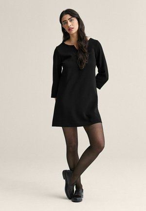 Kvinde iført en sort langærmet kjole, sorte gennemsigtige strømpebukser og sorte loafers, stående med krydsede ben mod en ensfarvet baggrund.