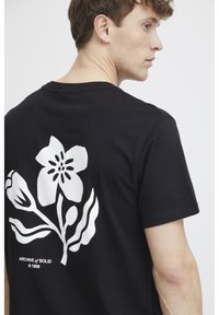 Sort bomulds T-shirt med et hvidt blomsterdesign på ryggen, med bladelementer og teksten "ARCHIVE of SOLID © 1999" nedenfor.
